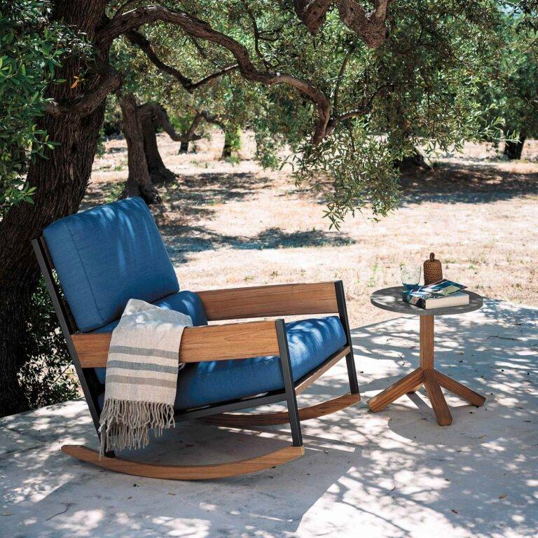 Ein Schaukelstuhl mit blauem Polster steht im Schatten eines Baumes.