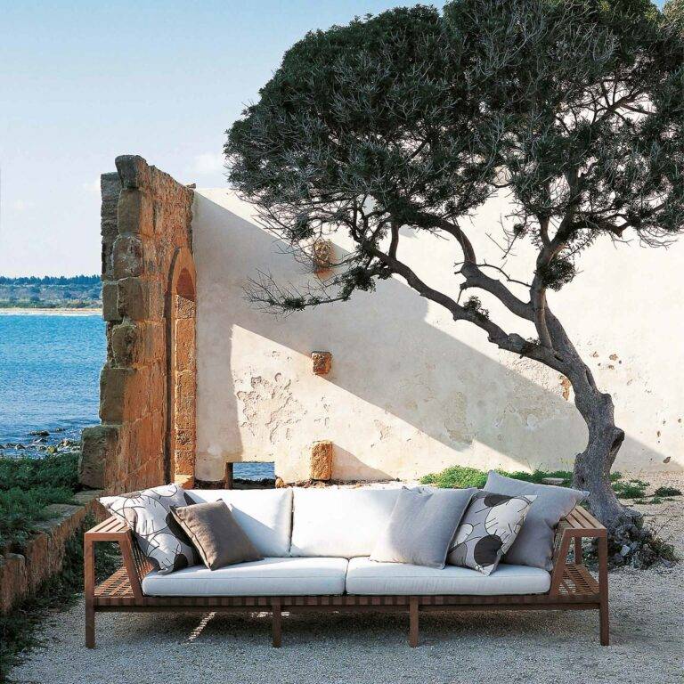 Eine Couch mit weißen Polstern steht an einer weißen Hauswand im Schatten eines Baumes mit dem Meer im Hintergrund.