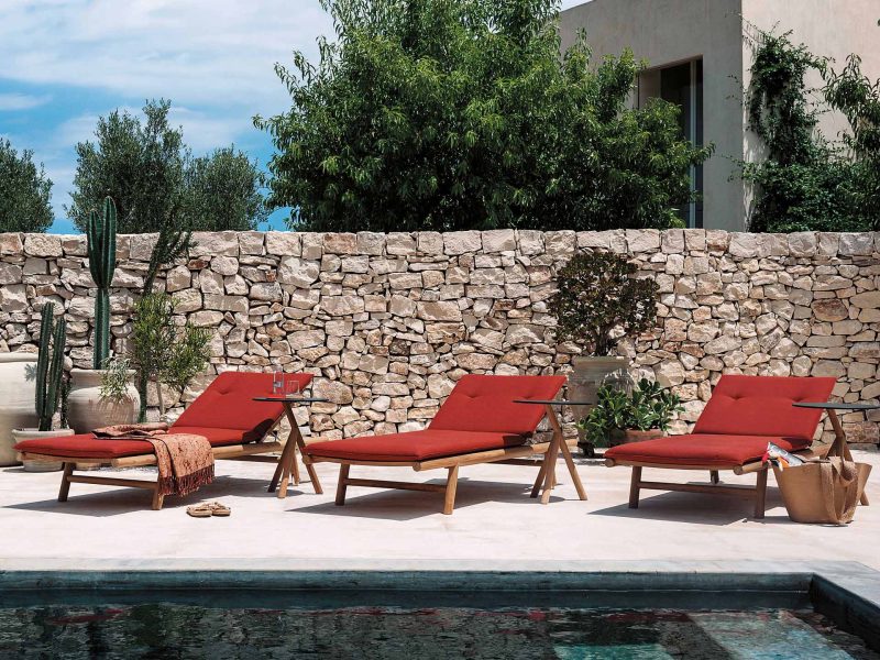 Drei Sonnenliegen mit roten Bezügen stehen in der Sonne an einem Pool.